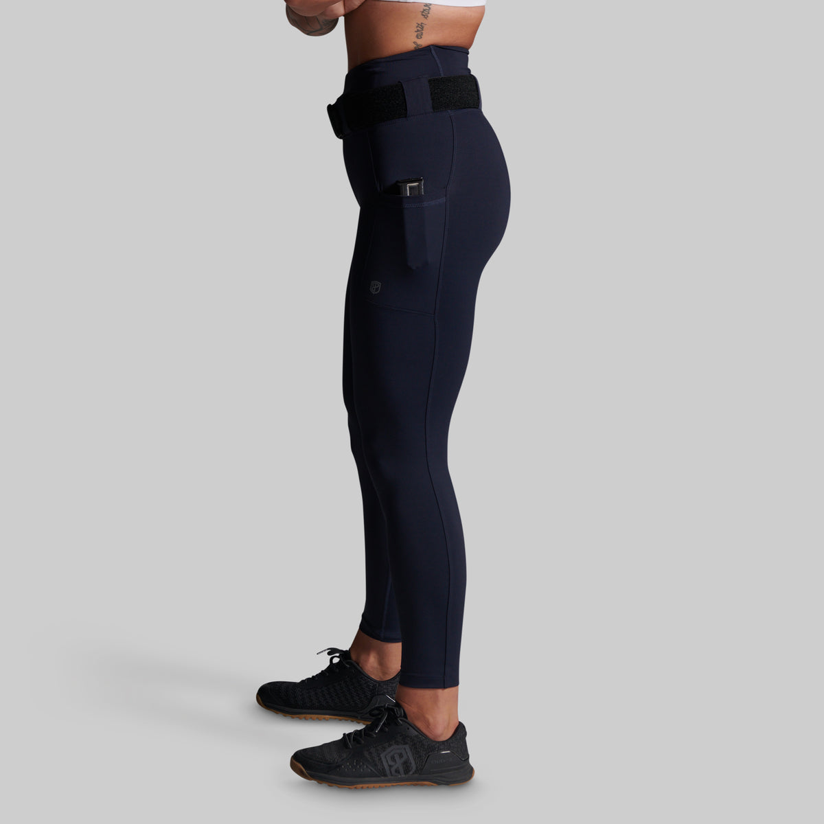 A side profile view of a person wearing dark navy blue athletic leggings with a side utility pocket and black shoes, set against a solid light gray background.