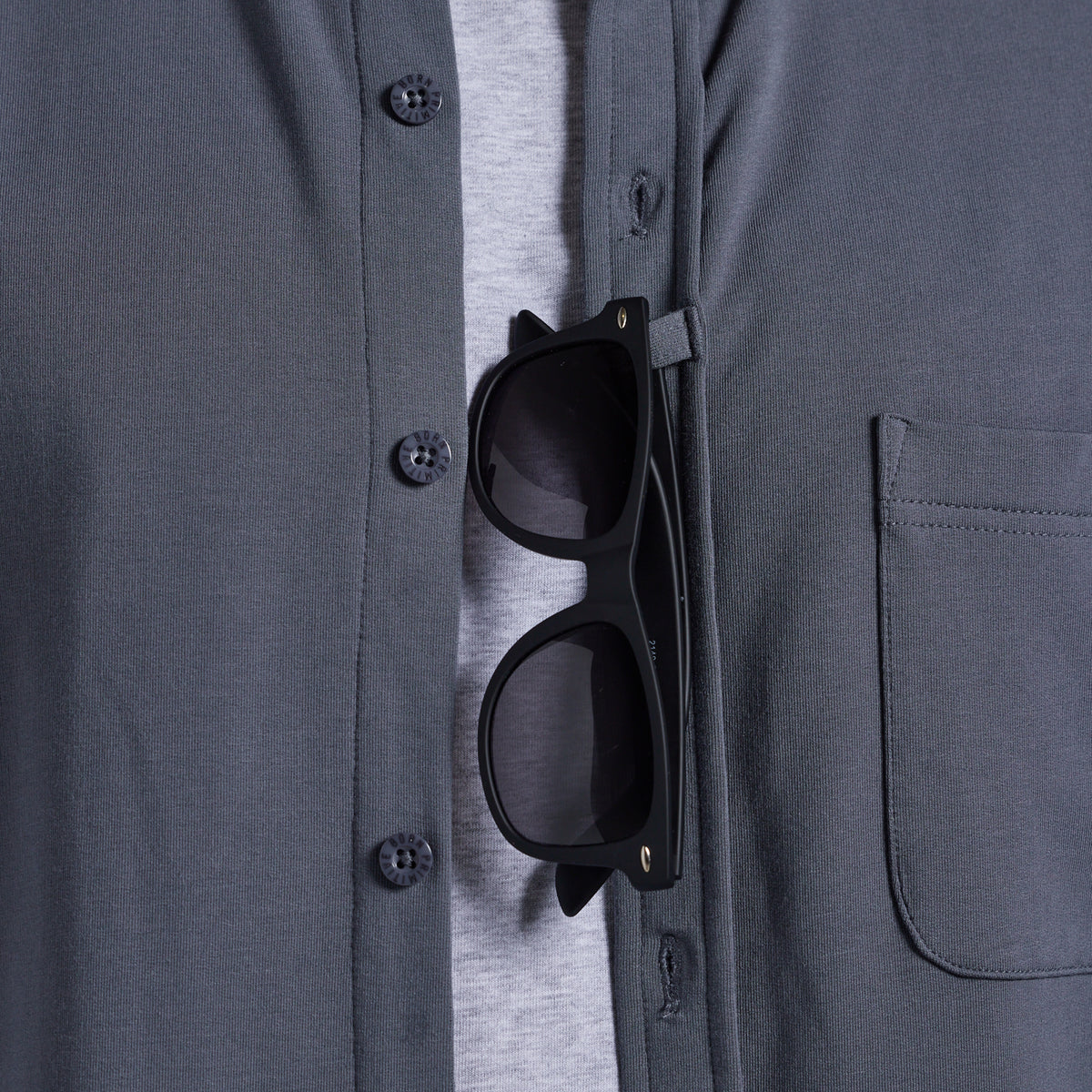Black sunglasses shown hanging from a pocket strap on a dark gray button-up shirt over a light gray t-shirt.