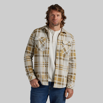 A man wearing a tan and cream plaid flannel shirt over a white henley tee and blue jeans, standing against a solid gray studio background.
