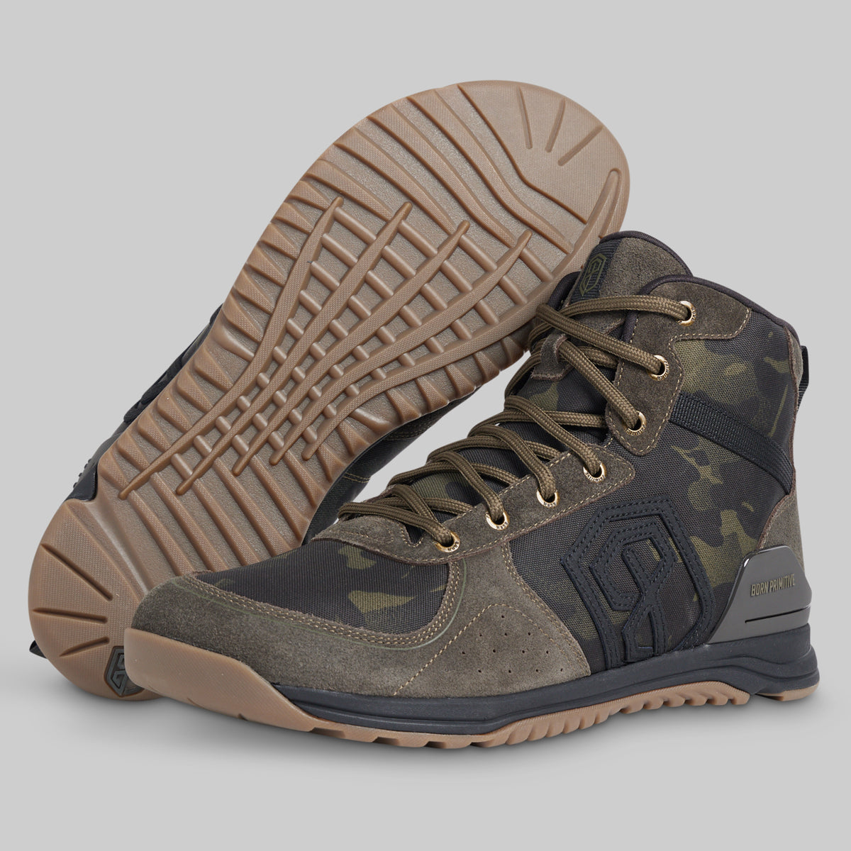 A pair of mid-cut tactical boots in a dark camouflage pattern with brown suede overlays and a tan outsole, shown against a solid gray background. One boot is positioned upright to show the side profile, while the other is turned to display the treaded outsole.