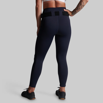 A model from the back wearing high-waisted navy blue activewear leggings with a black belt loop waistband, paired with black athletic shoes, against a solid gray studio background.