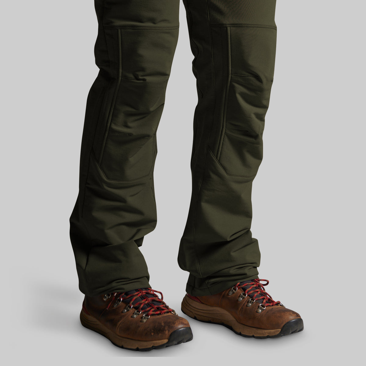 A close-up of a person's legs wearing dark olive green technical pants and brown hiking boots with red laces, shown against a solid gray background.