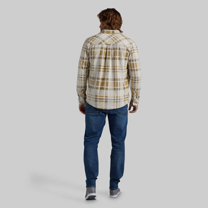 A man viewed from behind wearing a cream and tan plaid button-down shirt, blue jeans, and gray sneakers, standing against a solid light gray background.
