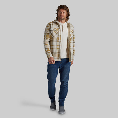 A man modeling a long-sleeve plaid flannel overshirt in beige and brown over a white henley shirt, paired with blue jeans and gray slip-on shoes, against a solid gray background.