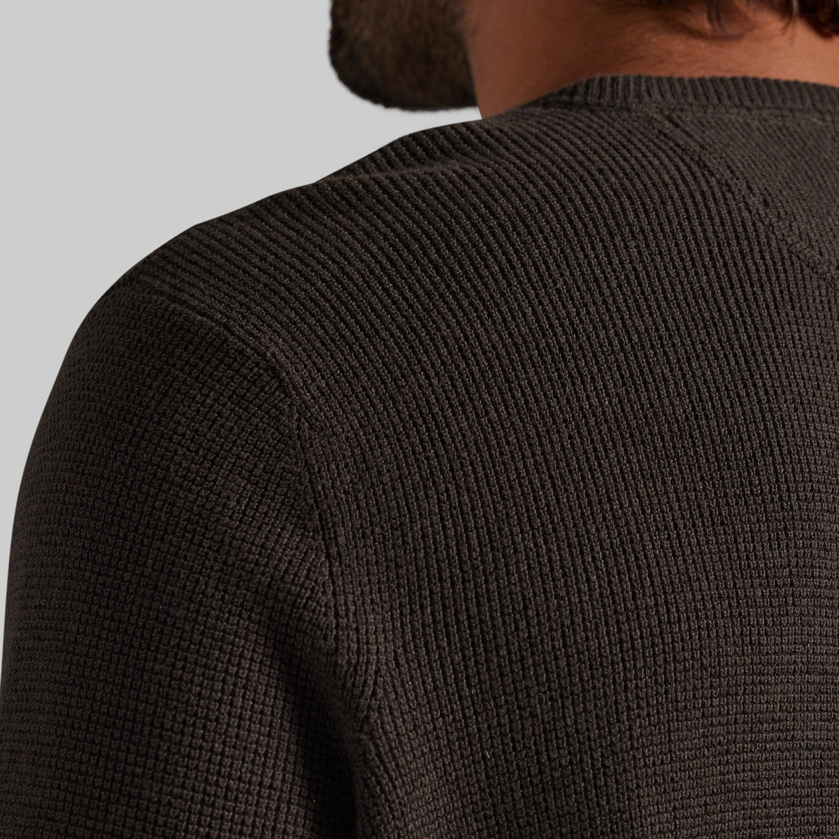 Close-up back view of a person wearing a dark brown ribbed knit sweater, showing the shoulder and upper back against a light gray background.