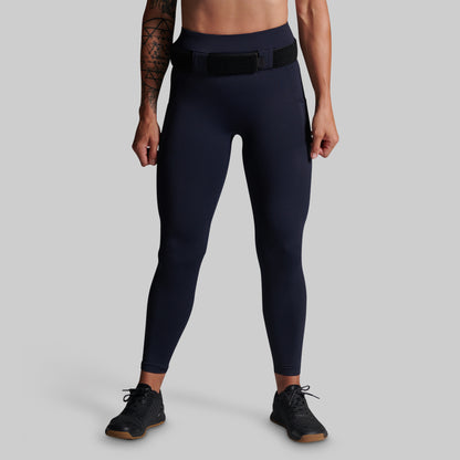 A model from the waist down wearing dark navy blue high-waisted athletic leggings with a black belt and black sneakers, set against a solid gray studio background.
