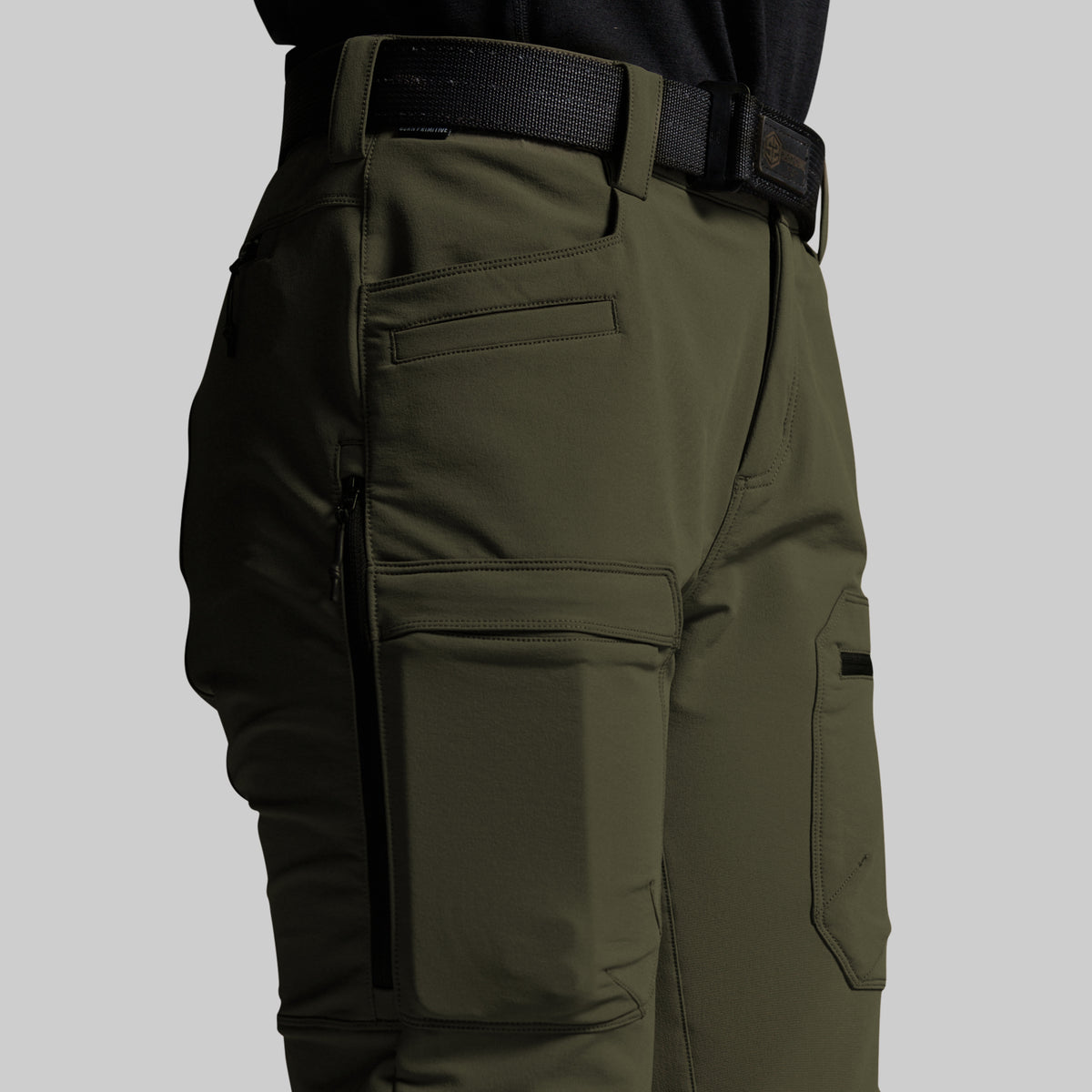Close-up of olive green outdoor cargo pants worn with a black belt, showing side pockets, zippered compartments, and a black top at the waist against a light gray background.