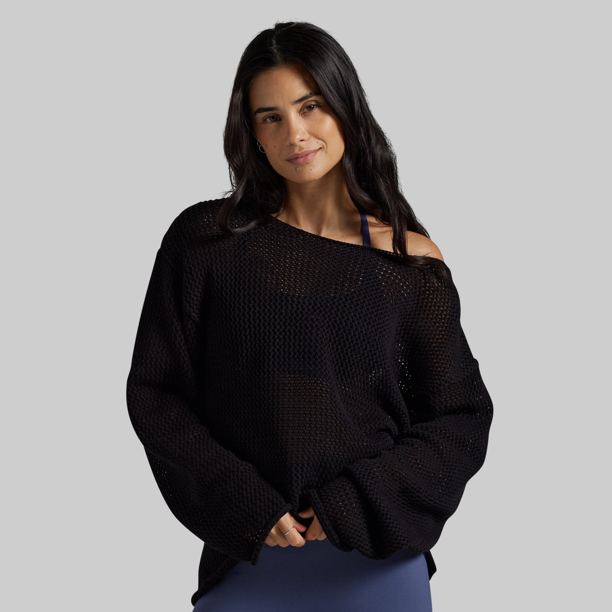 A woman modeling a black off-the-shoulder knit sweater with a textured open-knit pattern, paired with blue leggings, against a solid gray studio background.