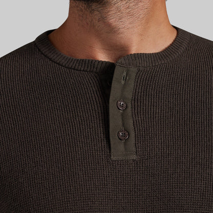 Close-up of a man wearing a dark brown ribbed knit henley shirt with a three-button placket on a light gray background.