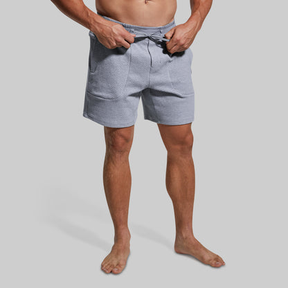 A man wearing light gray lounge shorts, shown from the waist down against a solid gray background.