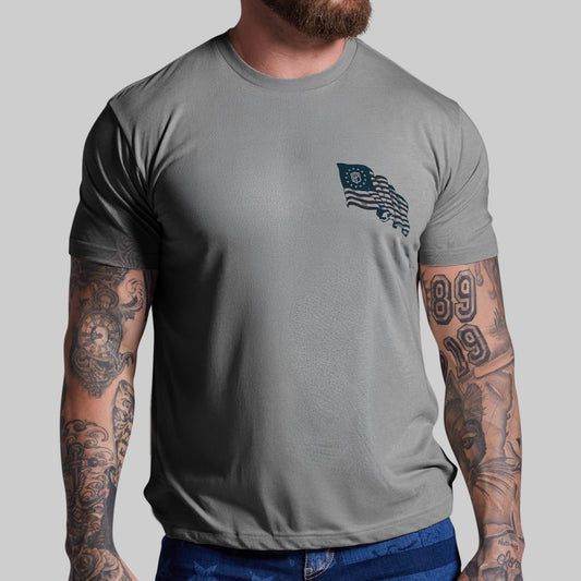 Man wearing a gray t-shirt with a small flag design on a plain background