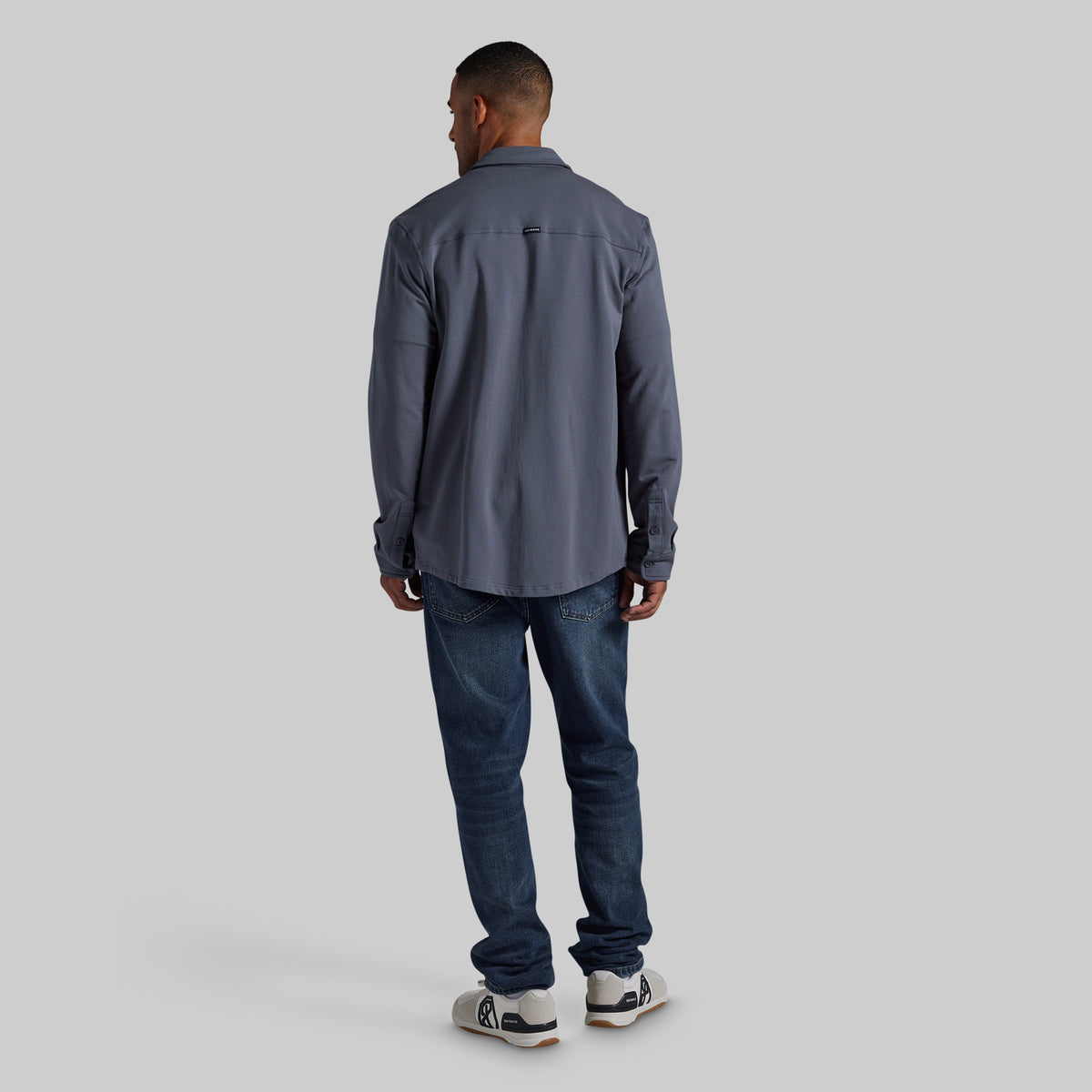 A man viewed from behind wearing a slate gray long-sleeve button-up shirt, dark blue jeans, and white sneakers, standing against a solid light gray background.