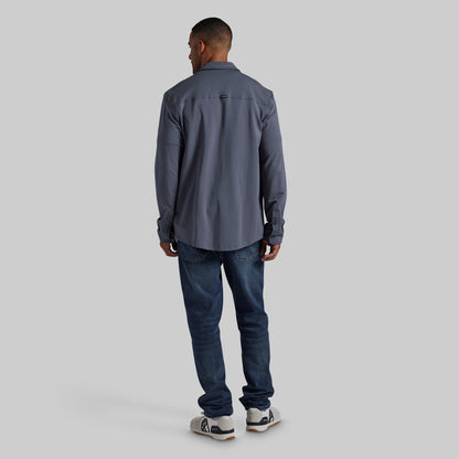 A man viewed from behind wearing a slate gray long-sleeve button-up shirt, dark blue jeans, and white sneakers, standing against a solid light gray background.
