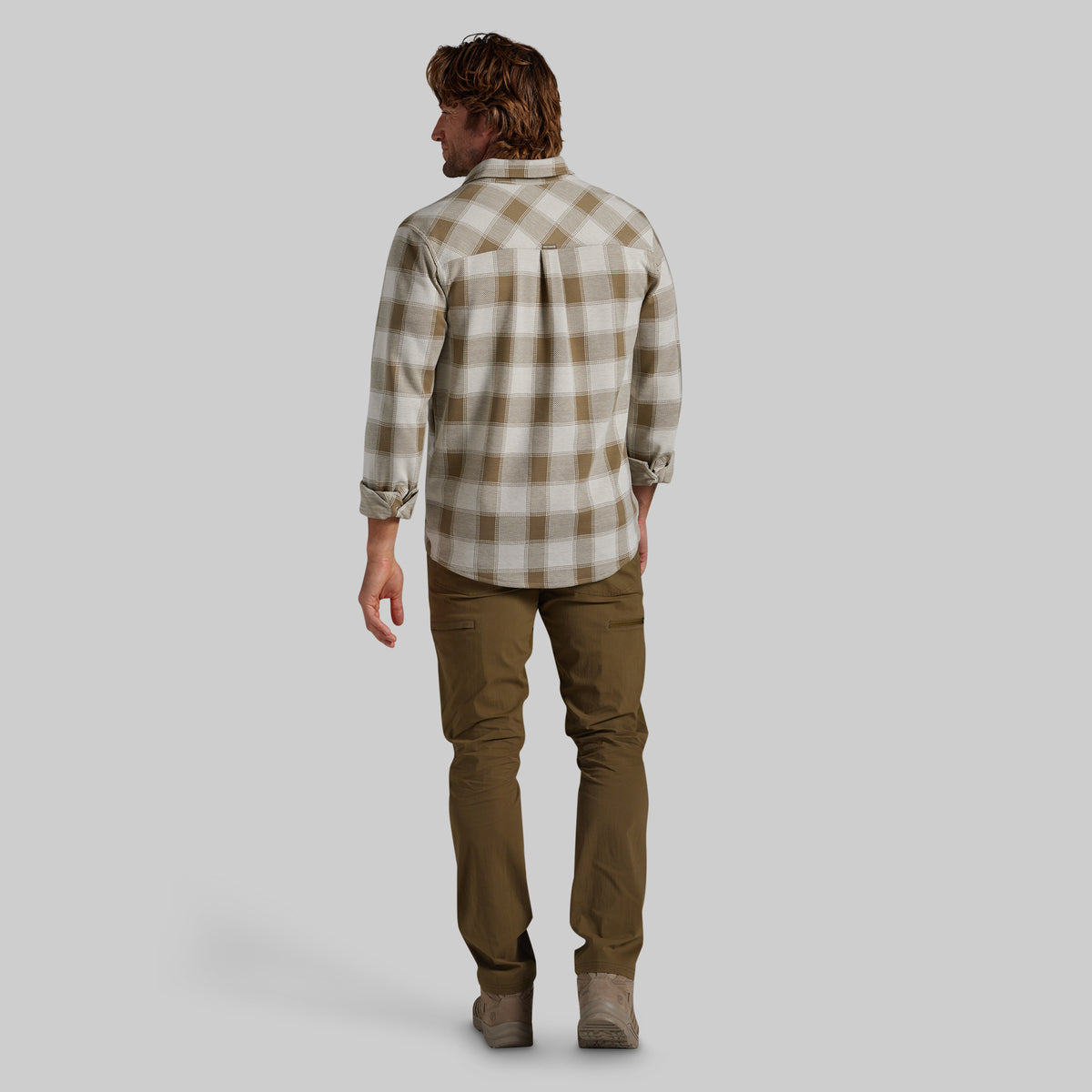 A back view of a man wearing a beige and brown plaid flannel shirt, paired with olive green pants, set against a solid gray background.