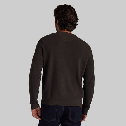 A man viewed from behind wearing a dark brown crewneck sweater and dark blue jeans, set against a solid light gray background.