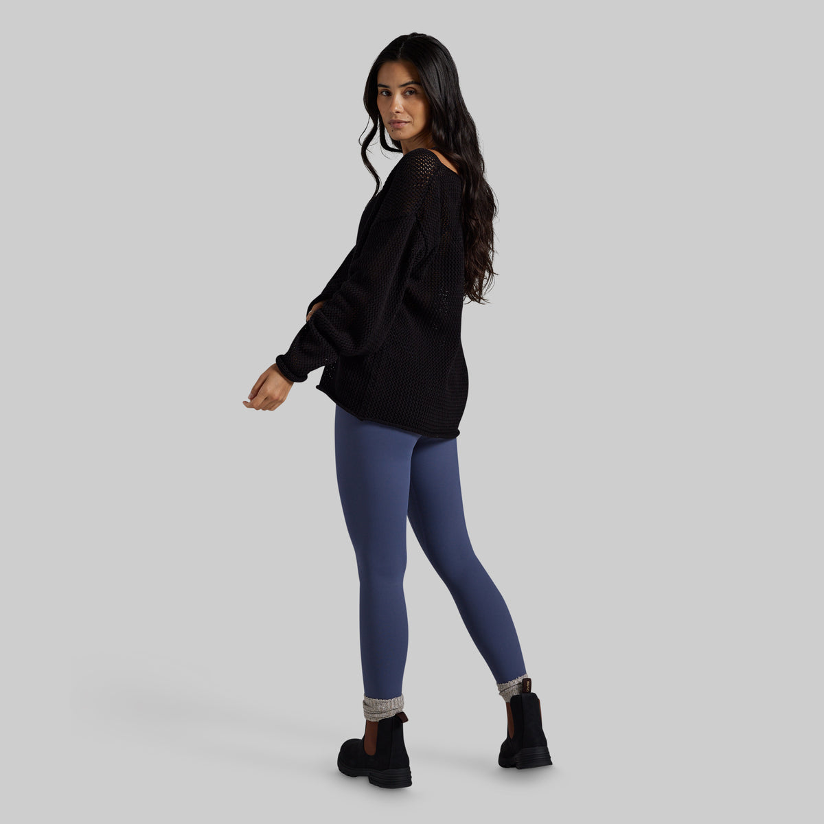 A woman modeling a black knit sweater and slate blue leggings with black ankle boots, shown from a three-quarters back view against a solid gray studio background.