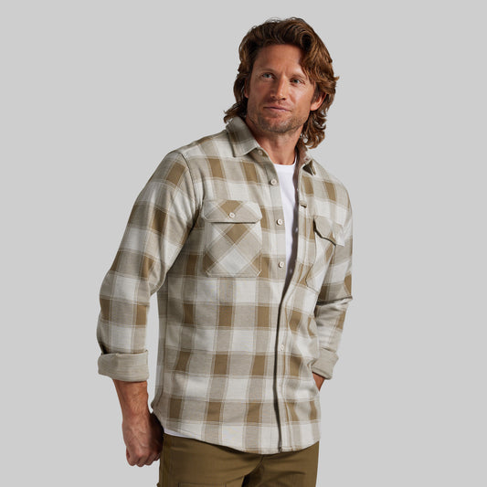 A man wearing a beige and brown plaid flannel shirt over a white undershirt, paired with olive green pants, set against a solid gray background.