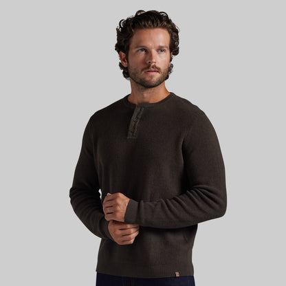 A man wearing a dark brown long-sleeve henley sweater, standing against a solid light gray background.