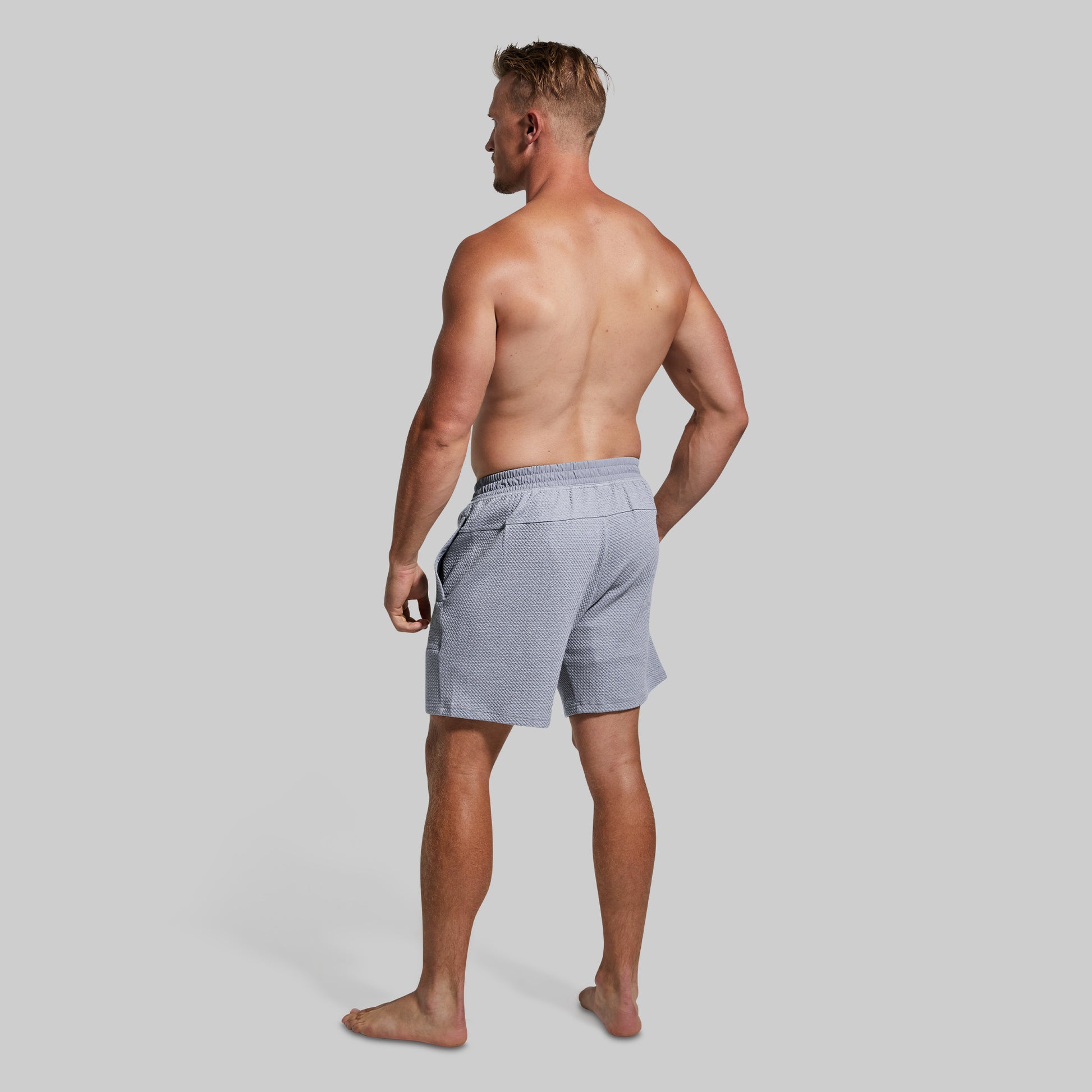 A shirtless man shown from the back wearing light gray textured lounge shorts, standing against a solid light gray studio background.