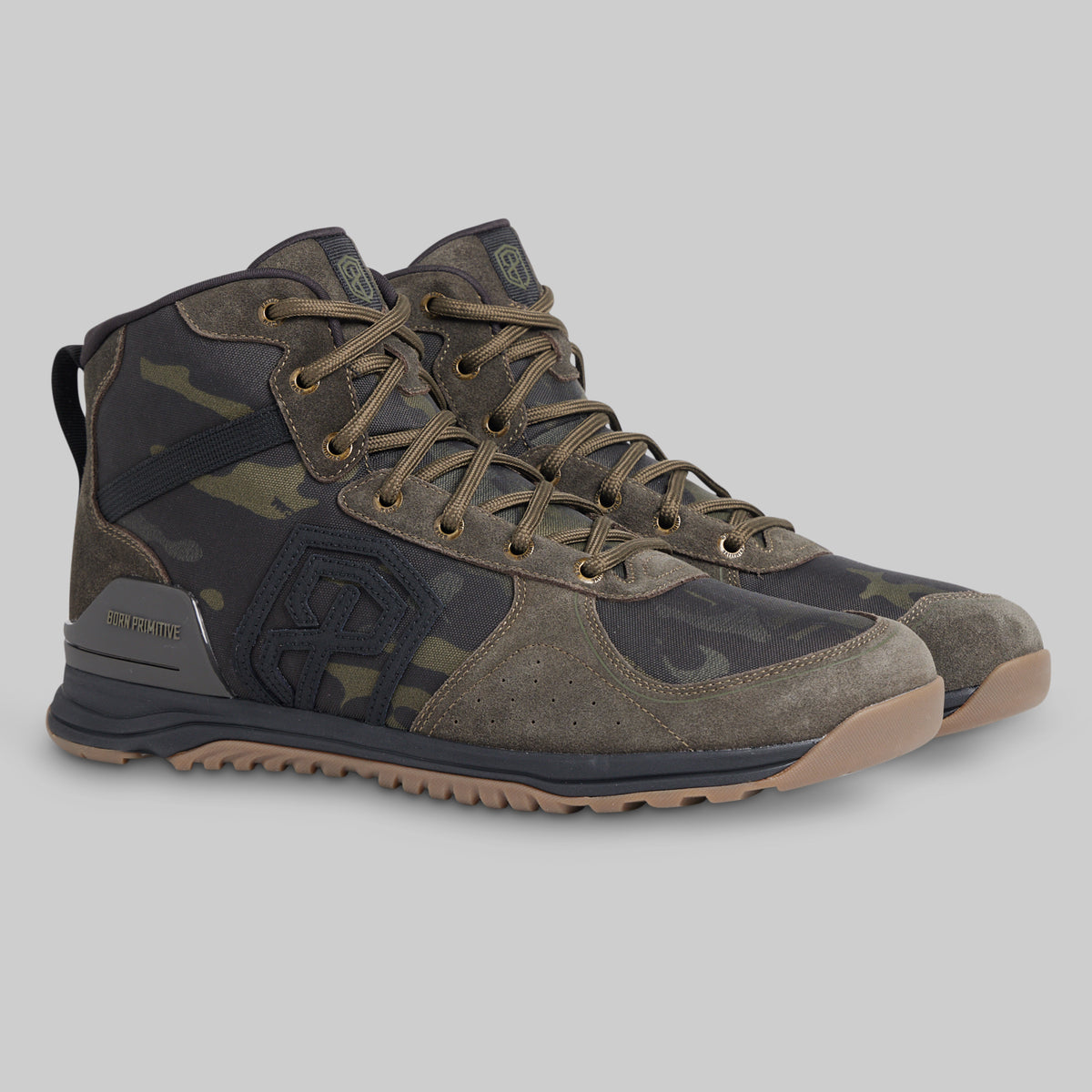 A pair of mid-top tactical boots in a dark camouflage pattern with olive green, black, and brown tones, featuring a rugged sole and lace-up closure, shown against a solid gray background.