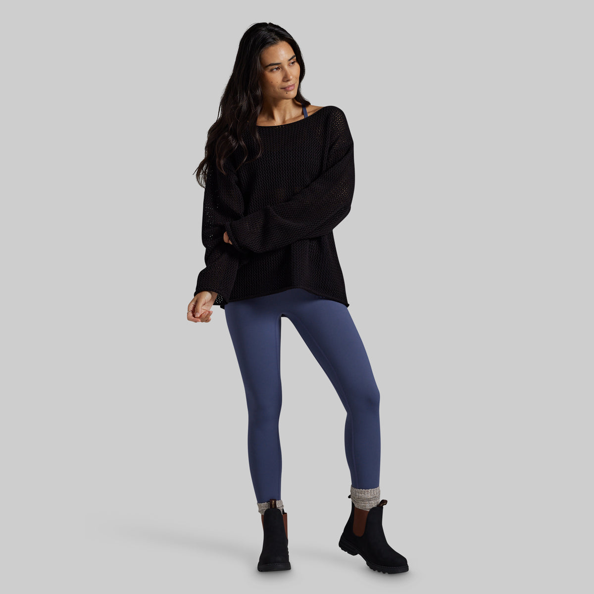 A woman modeling a black ribbed knit sweater with wide sleeves, paired with slate blue leggings and black ankle boots, against a solid gray studio background.