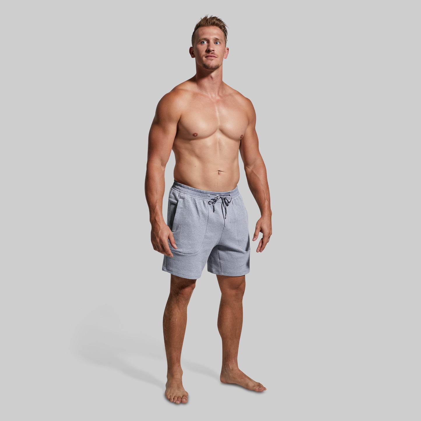A shirtless male model wearing light gray drawstring shorts, standing against a solid light gray studio background.
