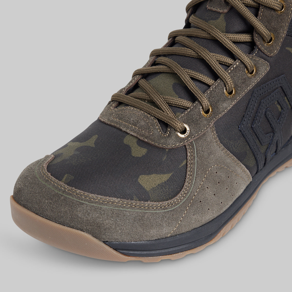 Close-up of a tactical boot featuring a dark green and black camouflage pattern, olive green suede overlays, matching laces, and a tan rubber outsole, set against a solid gray background.