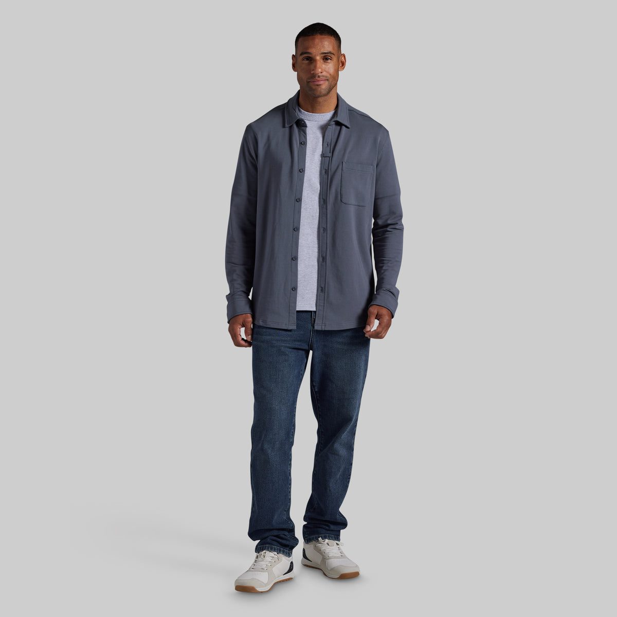 A man modeling a slate gray button-down shirt over a white t-shirt, paired with dark blue jeans and white sneakers, standing against a solid gray background.