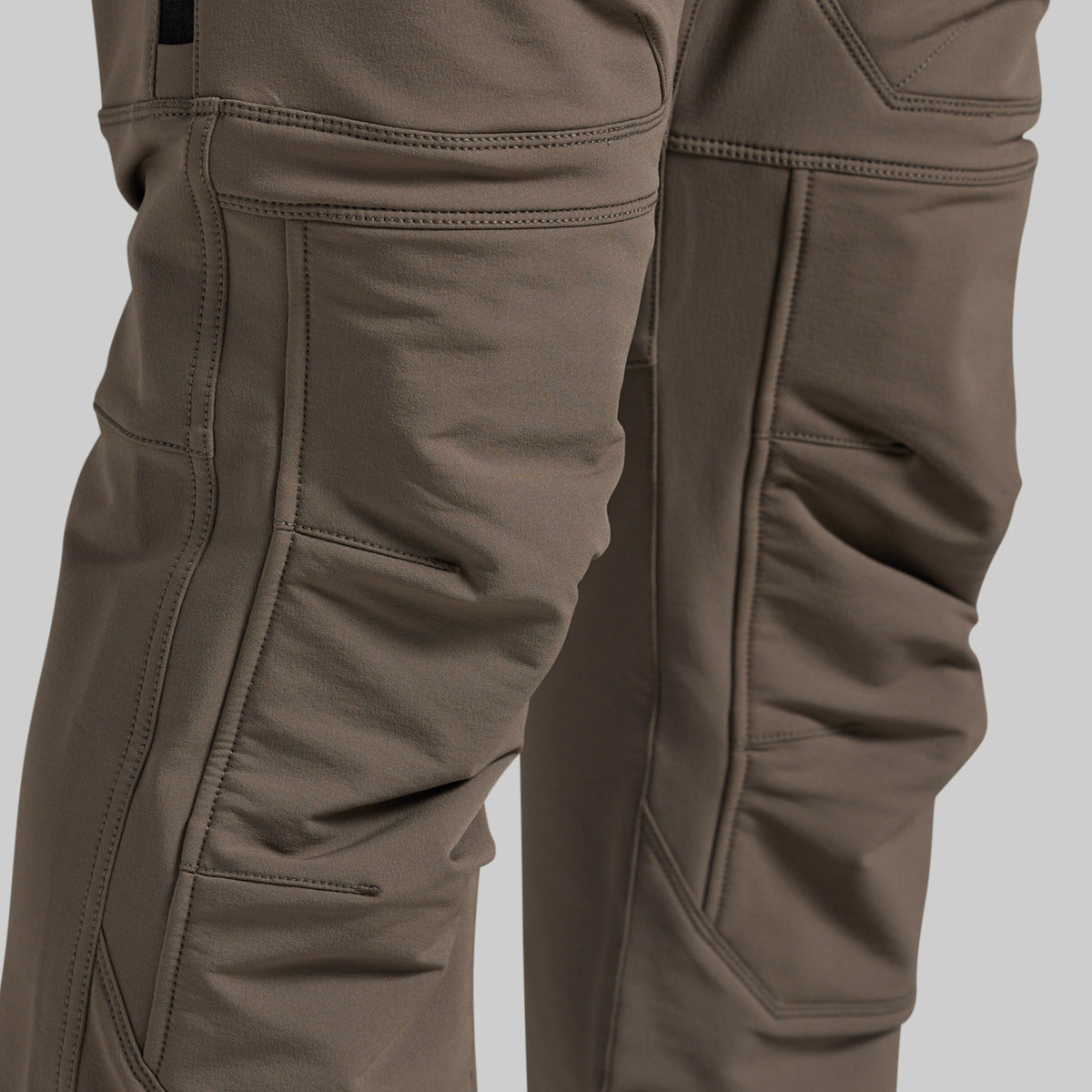 Close-up of a pair of technical outdoor pants in a warm grey color, showing the detailed stitching, reinforced knees, and fabric texture.