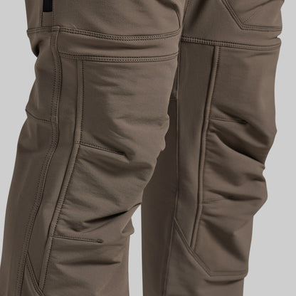 Close-up of a pair of technical outdoor pants in a warm grey color, showing the detailed stitching, reinforced knees, and fabric texture.