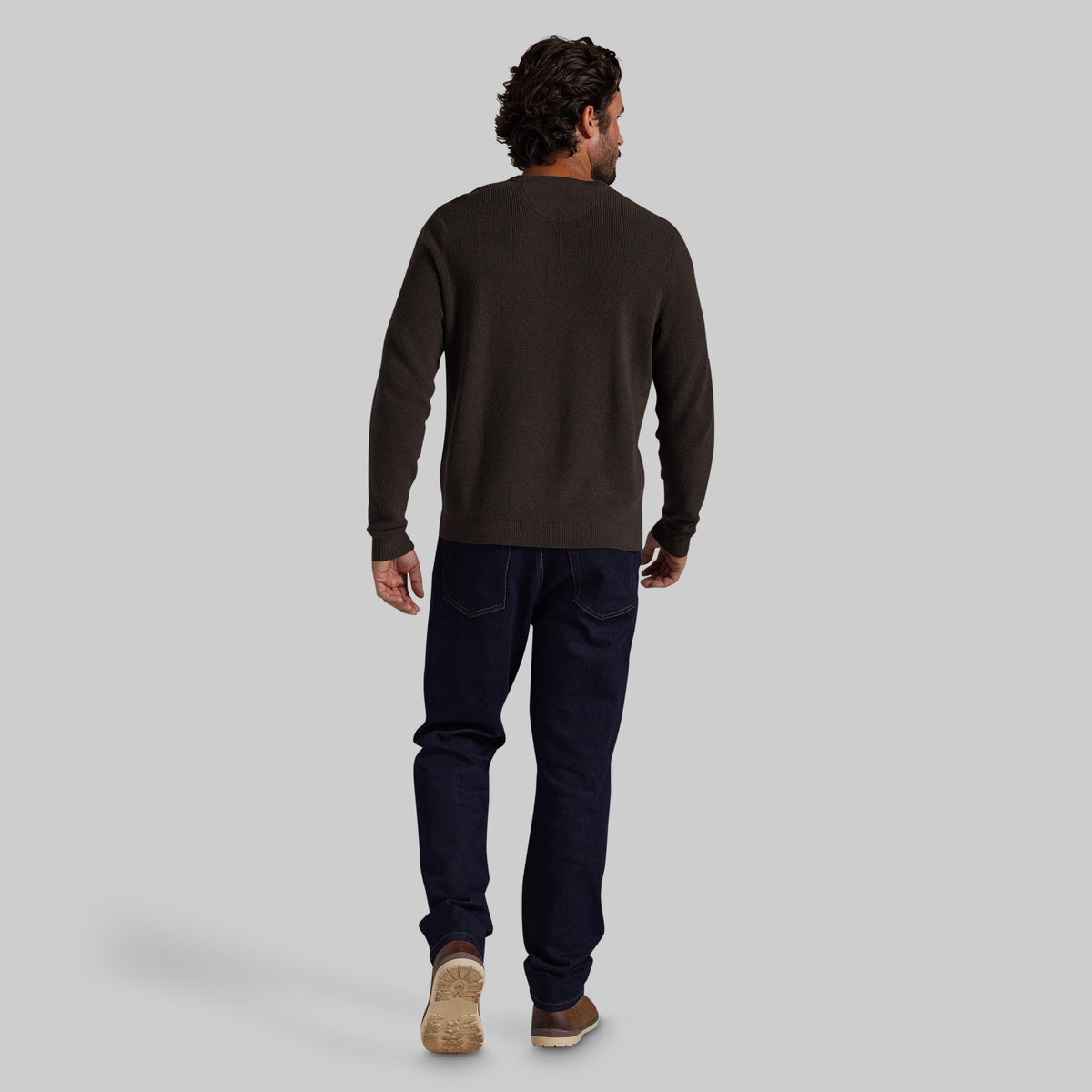 A man is shown from behind wearing a dark brown long-sleeve crewneck sweater, dark navy blue jeans, and brown shoes, standing against a solid light gray background.
