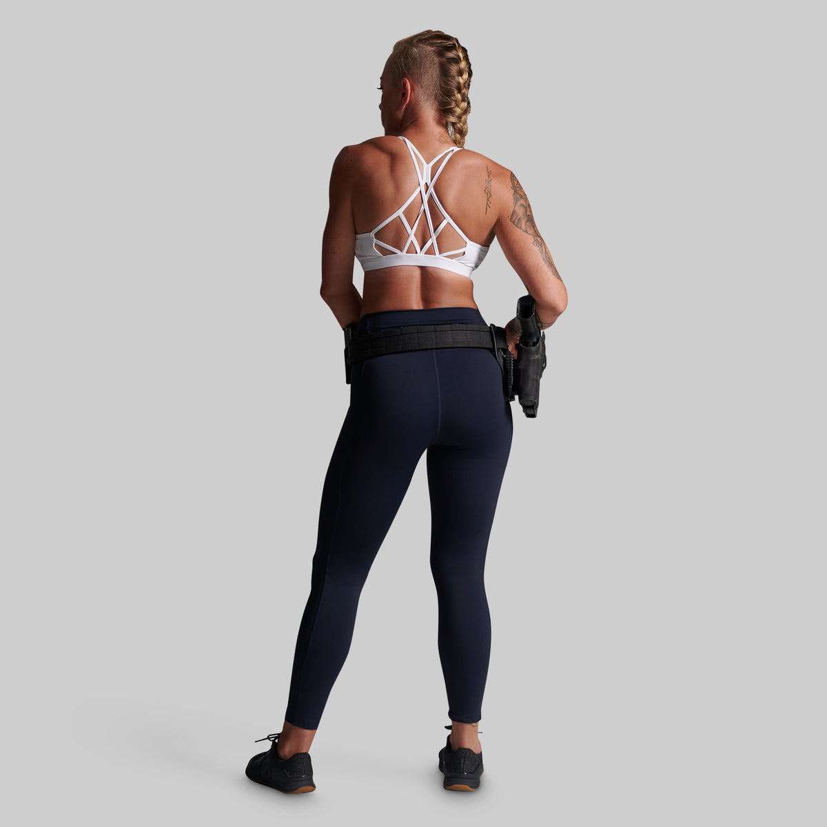 A woman shown from the back wearing a white strappy sports bra and navy blue leggings, standing against a light gray studio background.