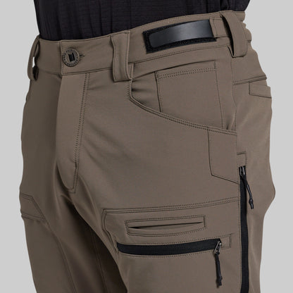 Close-up of a person wearing outdoor cargo pants in a storm grey color, featuring a black belt, belt buckle, and multiple zippered pockets.