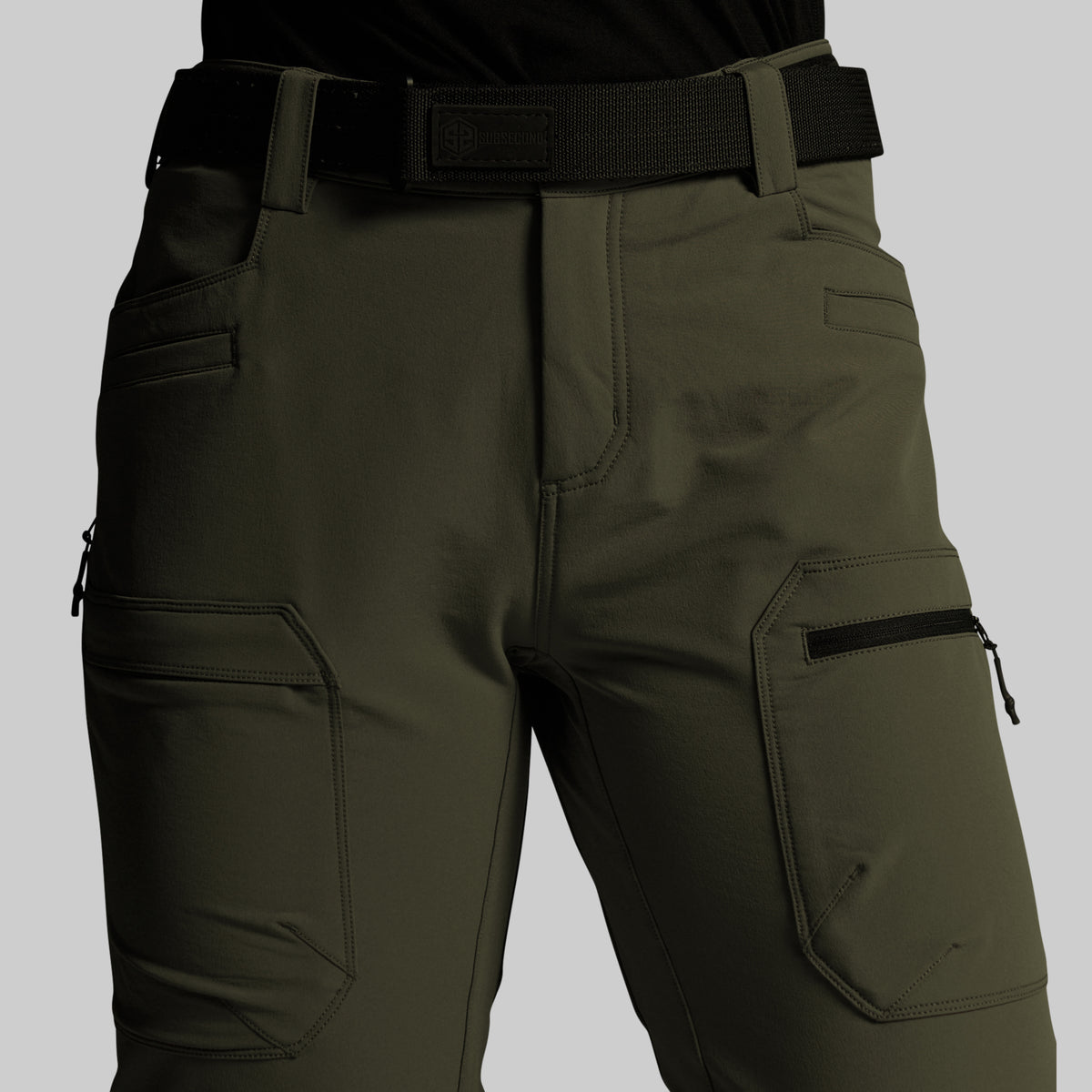 Olive green outdoor cargo pants with black belt and black zipper details, shown on a model from the waist down against a solid gray background.