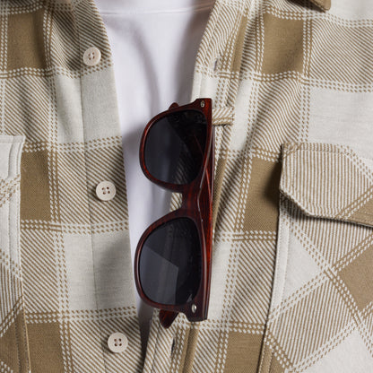 A close-up of a person wearing a beige and white plaid flannel shirt over a white undershirt, with dark brown sunglasses hanging from the placket.