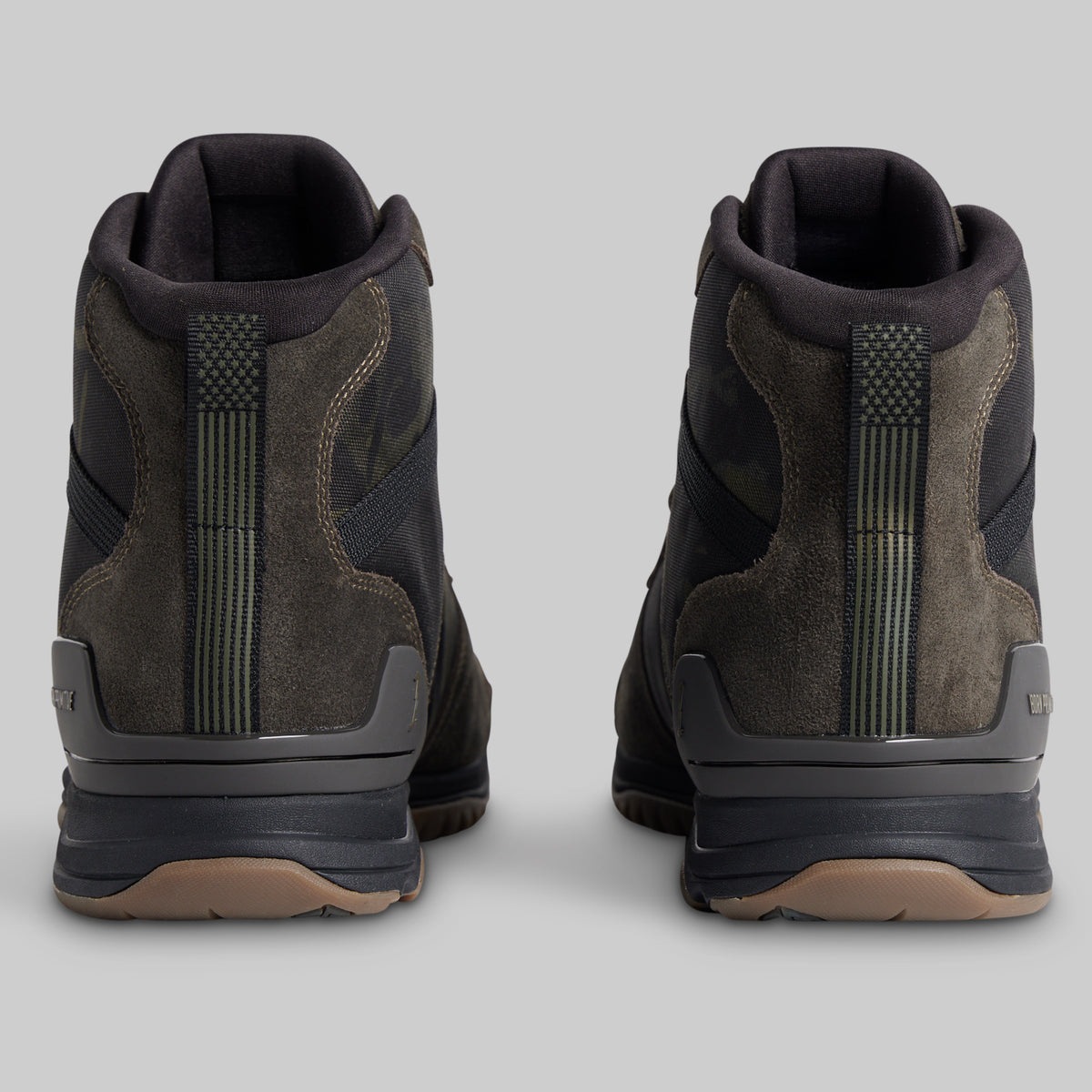 A pair of mid-height tactical boots shown from the back, featuring dark brown suede and black synthetic panels with olive green details, set against a solid gray background.