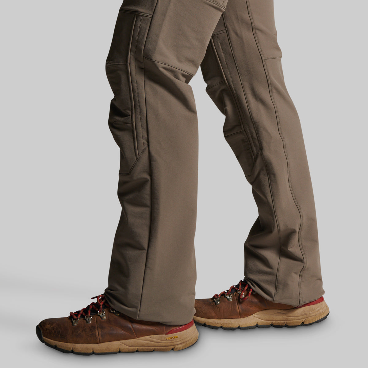 Close-up of a person's legs wearing brown hiking boots and grey pants, shown from the knees down against a light gray studio background.