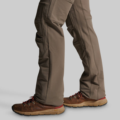 Close-up of a person's legs wearing brown hiking boots and grey pants, shown from the knees down against a light gray studio background.