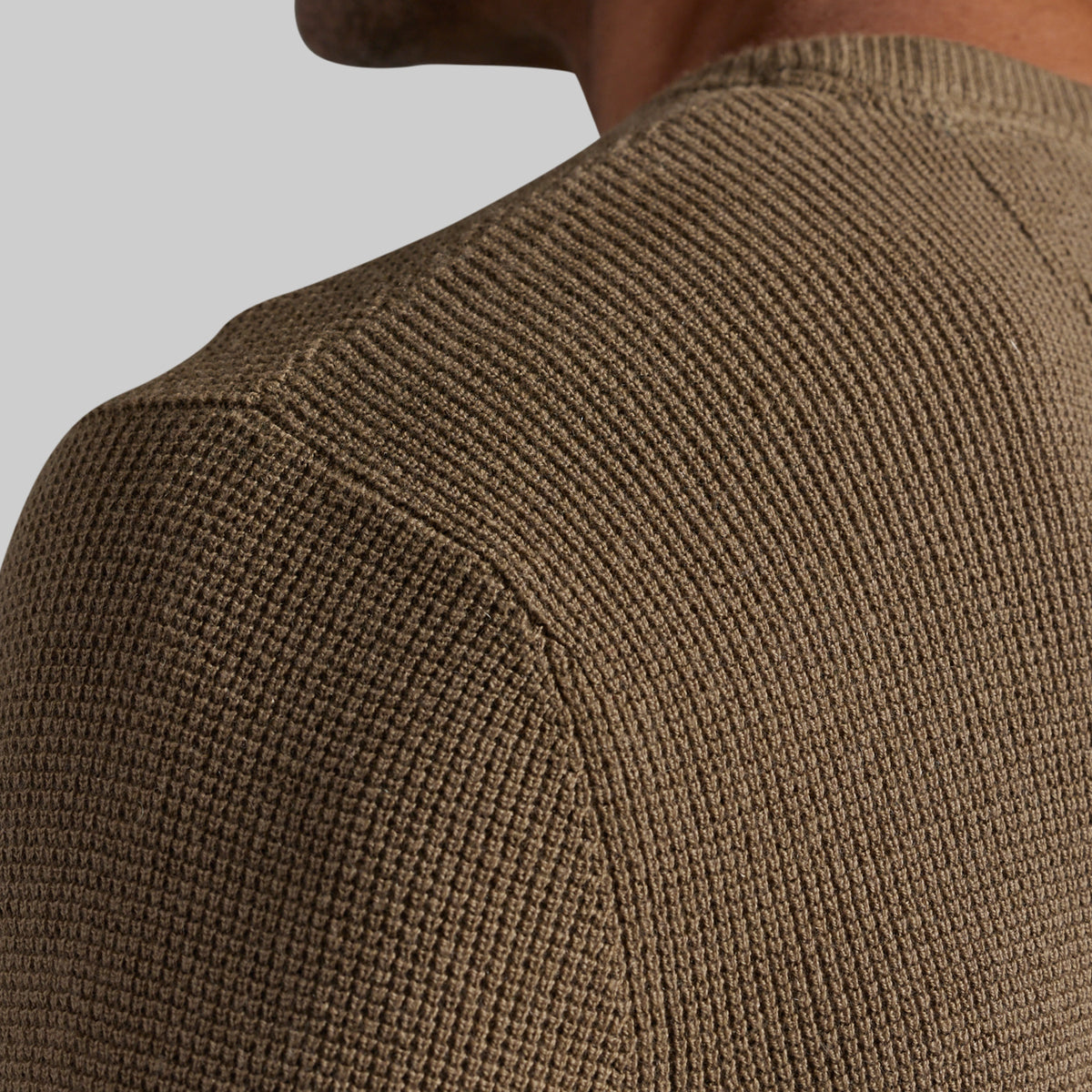 Close-up of a person wearing a ribbed knit sweater, showing the shoulder and upper chest area against a plain light gray background.