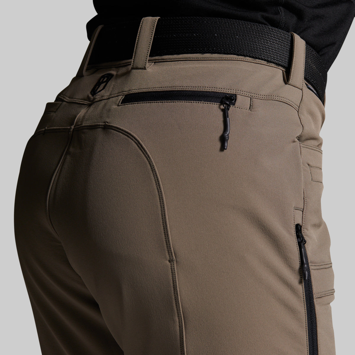 Close-up of the back waist and pockets of grey outdoor pants, featuring a black belt, a zippered pocket, and a vertical cargo pocket on a light gray background.