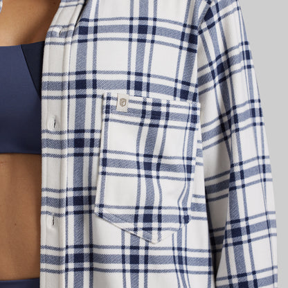 Close-up of a person wearing a white button-up shirt with a navy blue plaid pattern, featuring a chest pocket with a small brand tag.