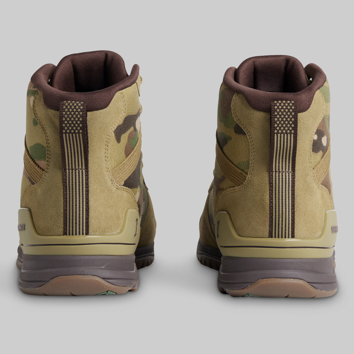 A pair of mid-cut tactical boots shown from the back, featuring a tan and green camouflage pattern with brown accents, set against a solid gray background.