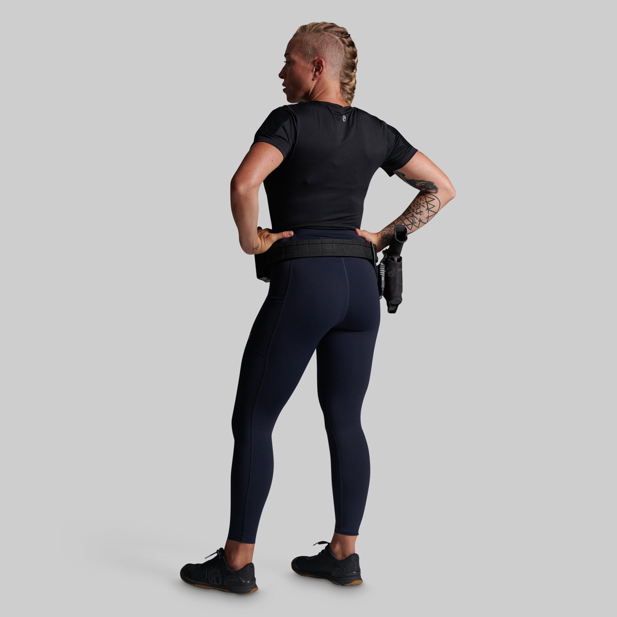 A woman shown from the back wearing dark navy leggings, a black short-sleeve top, and black sneakers, with a black tactical belt.