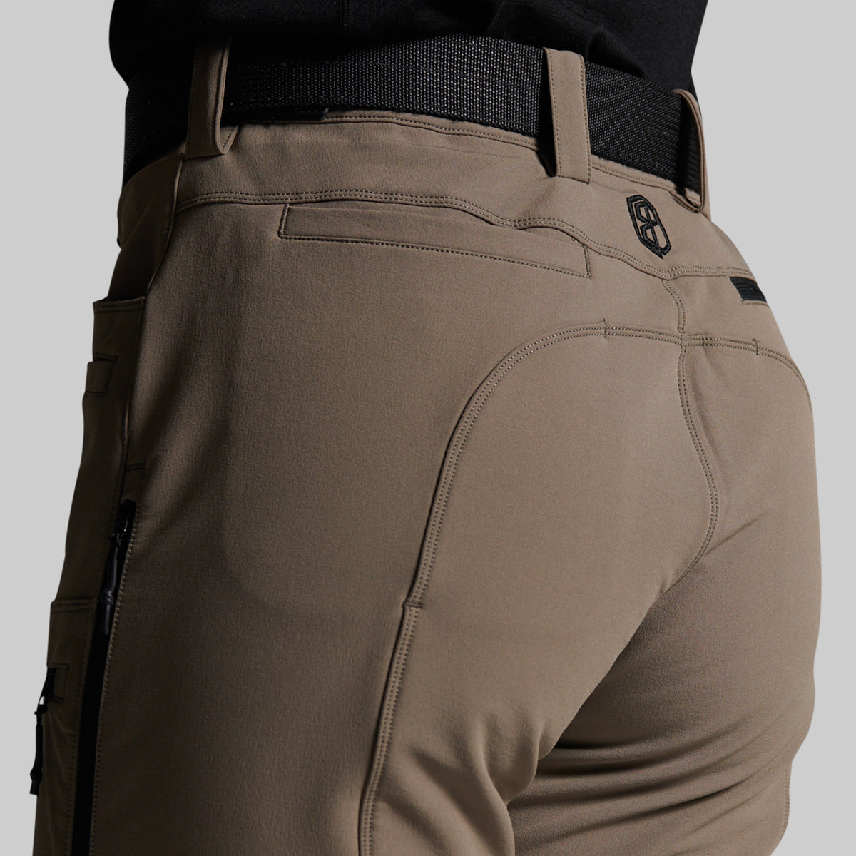 Close-up rear view of a person wearing grey outdoor cargo pants with a black belt, showing the back pocket and hip design against a light gray studio background.