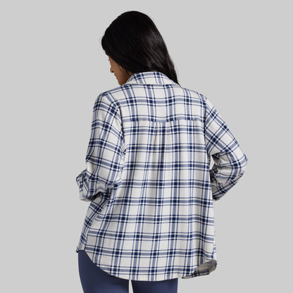 A woman shown from the back wearing a white long-sleeve plaid button-up shirt with navy and thin gray stripes, paired with dark blue jeans, against a solid gray background.