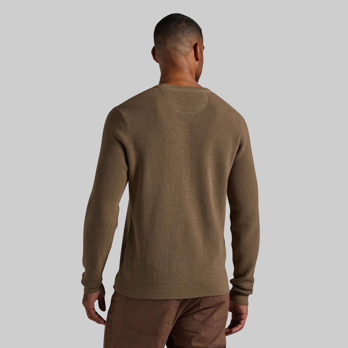 A man viewed from the back wearing a brown knit crewneck sweater and brown pants against a solid light gray background.