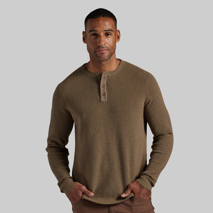 A man wearing a brown ribbed knit long-sleeve henley shirt with a three-button placket, standing against a solid gray background.
