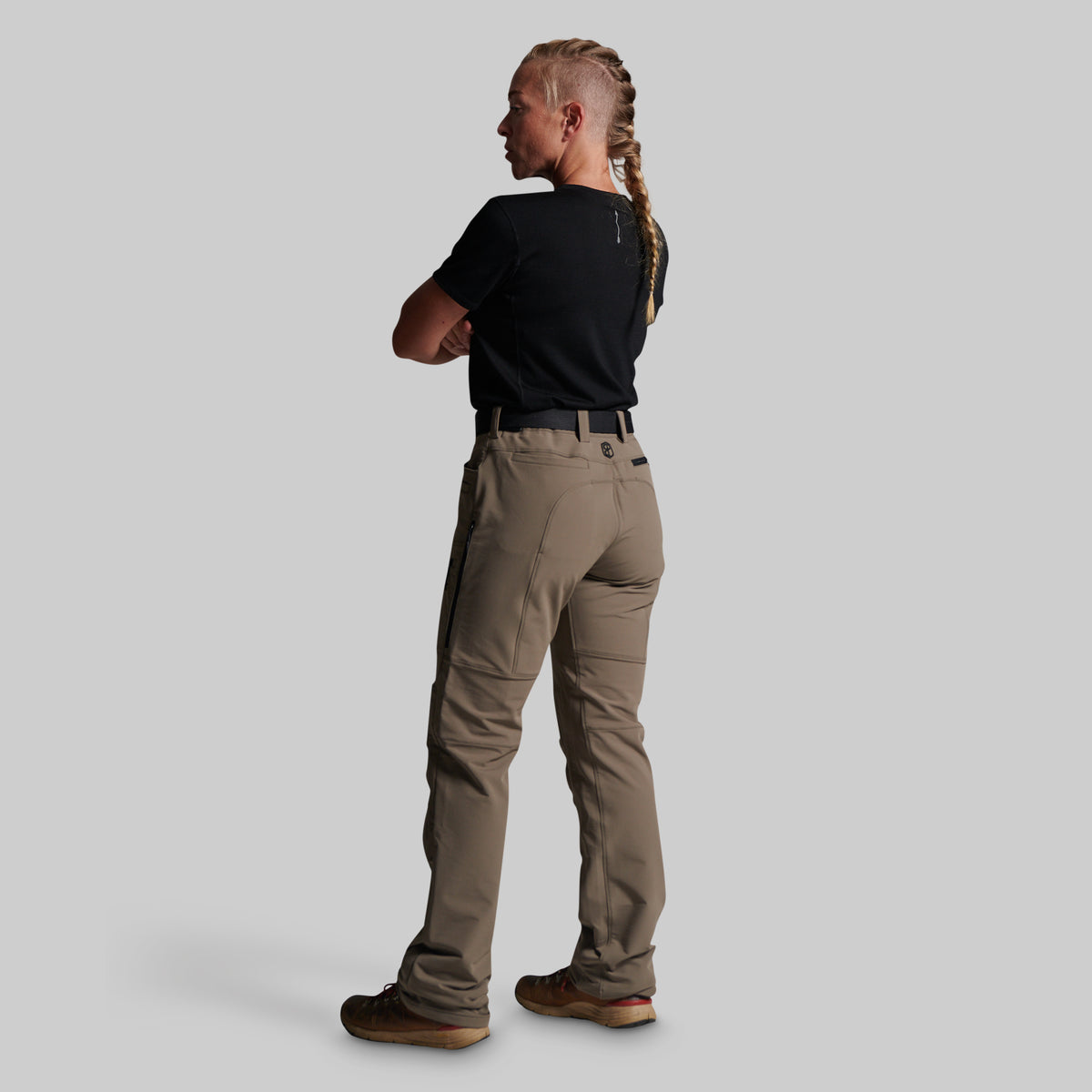 A woman shown from the back wearing grey cargo-style pants, a black short-sleeve top, and brown shoes against a plain light gray studio background.