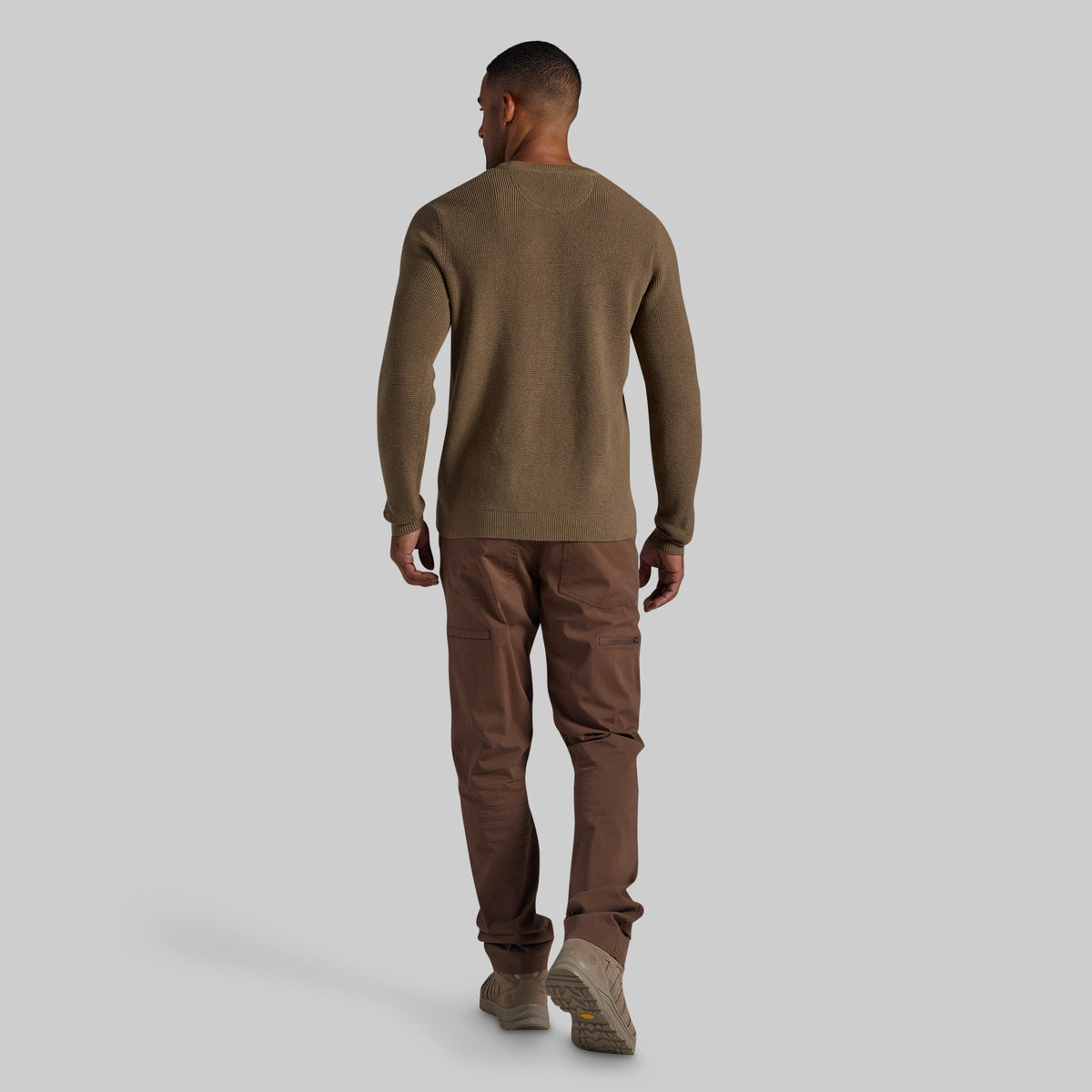 A man shown from the back wearing a brown long-sleeve knit sweater, brown pants, and brown shoes, standing against a solid light gray background.