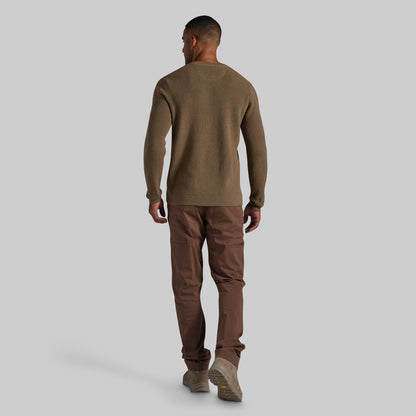 A man shown from the back wearing a brown long-sleeve knit sweater, brown pants, and brown shoes, standing against a solid light gray background.